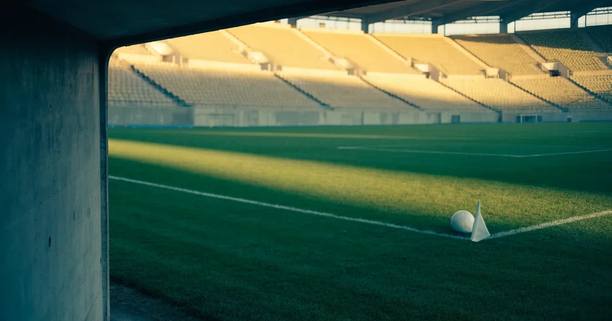 Blick durch ein Stadiontor auf einen grünen Rasen im Abendlicht, ein Ball ruht an der Eckfahne, die Tribünen sind leer.