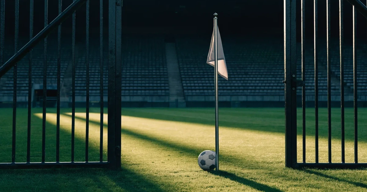 Blick durch ein Stadiontor auf einen grünen Rasen im Abendlicht, ein Ball liegt an der Eckfahne, die Ränge sind leer.