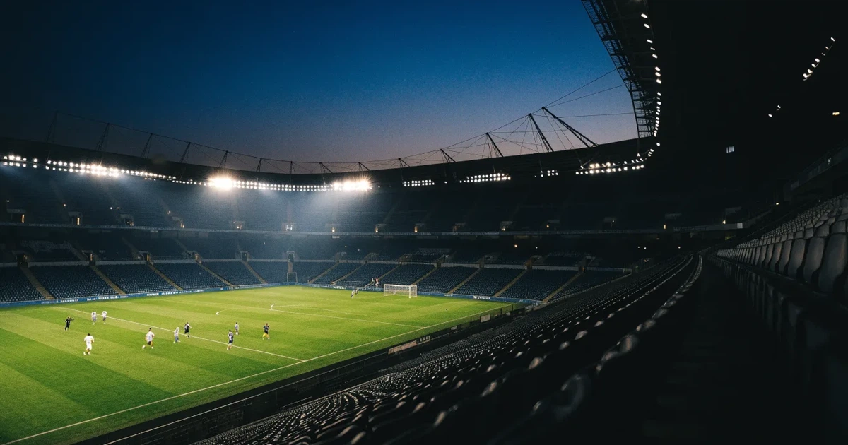 Leere Sitzreihen eines großen Stadions unter Flutlicht, dahinter die ferne Sicht auf den hell beleuchteten Rasen nach Sonnenuntergang.