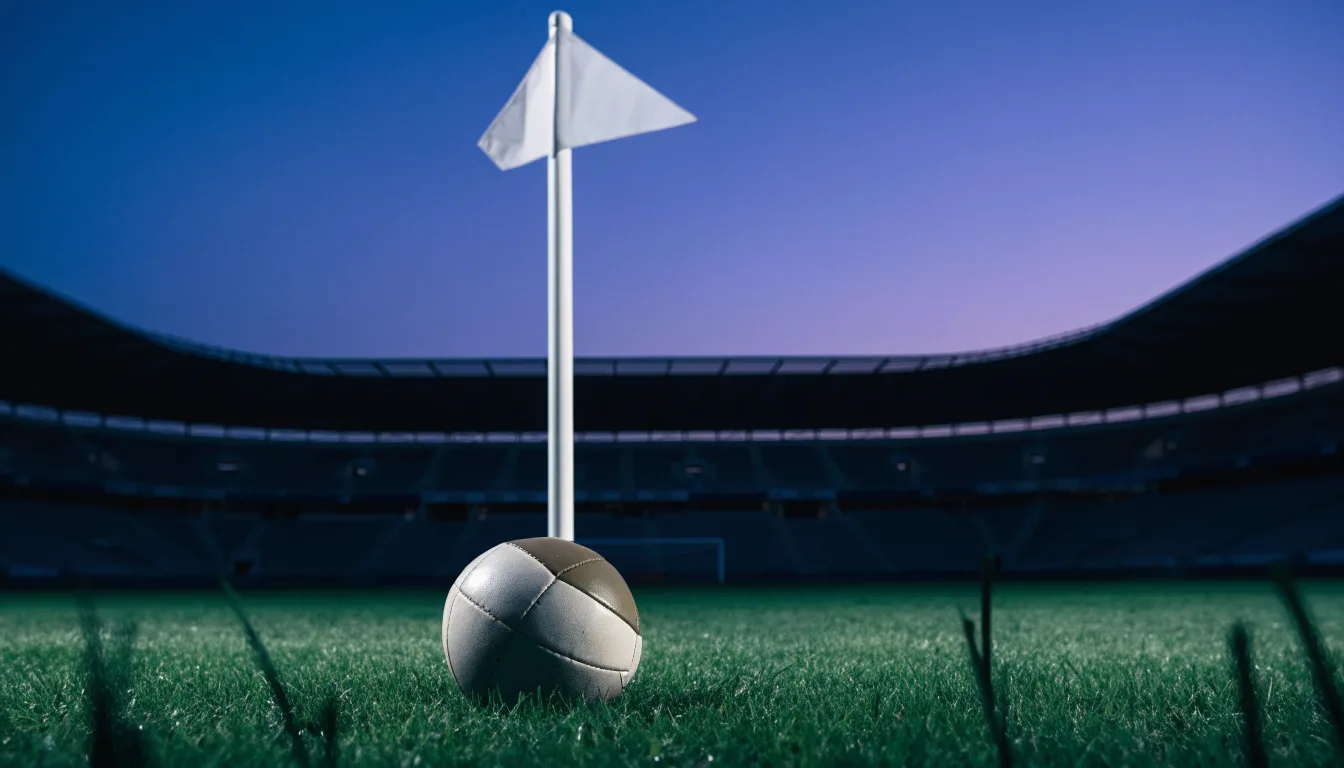 Ein Fußball liegt neben einem Eckfahnen-Pfosten auf einem Rasenplatz, im Hintergrund ein leeres Stadion in der Dämmerung.