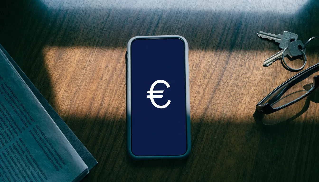 Ein Smartphone liegt mit dunklem Display auf einem Holztisch neben einem Schlüsselbund und einer Brille im Licht eines Fensters.