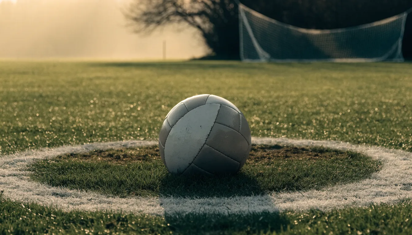 Ein Fußball liegt auf dem Anstoßkreis eines leeren Rasenplatzes im Licht der Morgensonne.