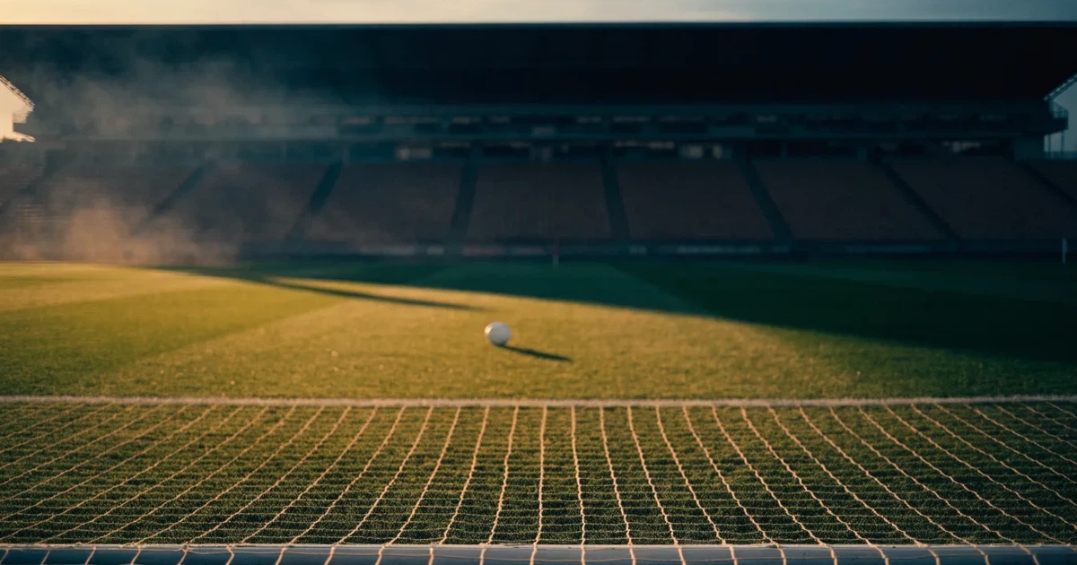 Ein Fußballplatz mit frisch gemähtem Rasen im Abendlicht, ein Ball liegt im Strafraum, im Hintergrund unscharf eine leere Tribüne.