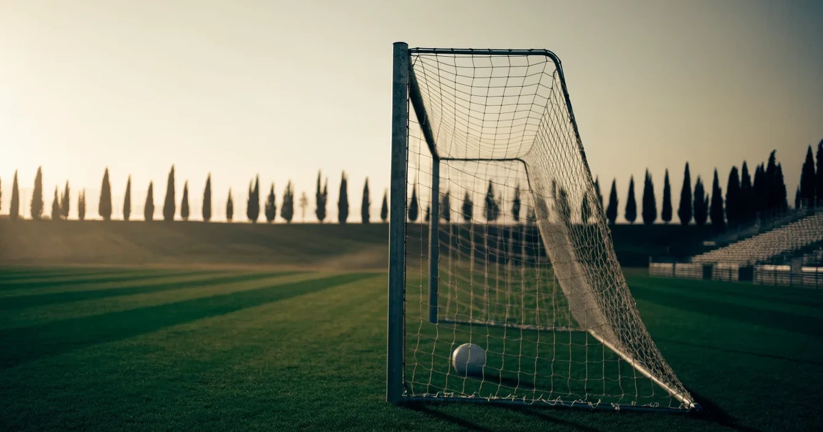Ein Fußballtor mit Netz auf einem leeren Platz im Abendlicht, der Rasen frisch gemäht, im Hintergrund Zypressen.