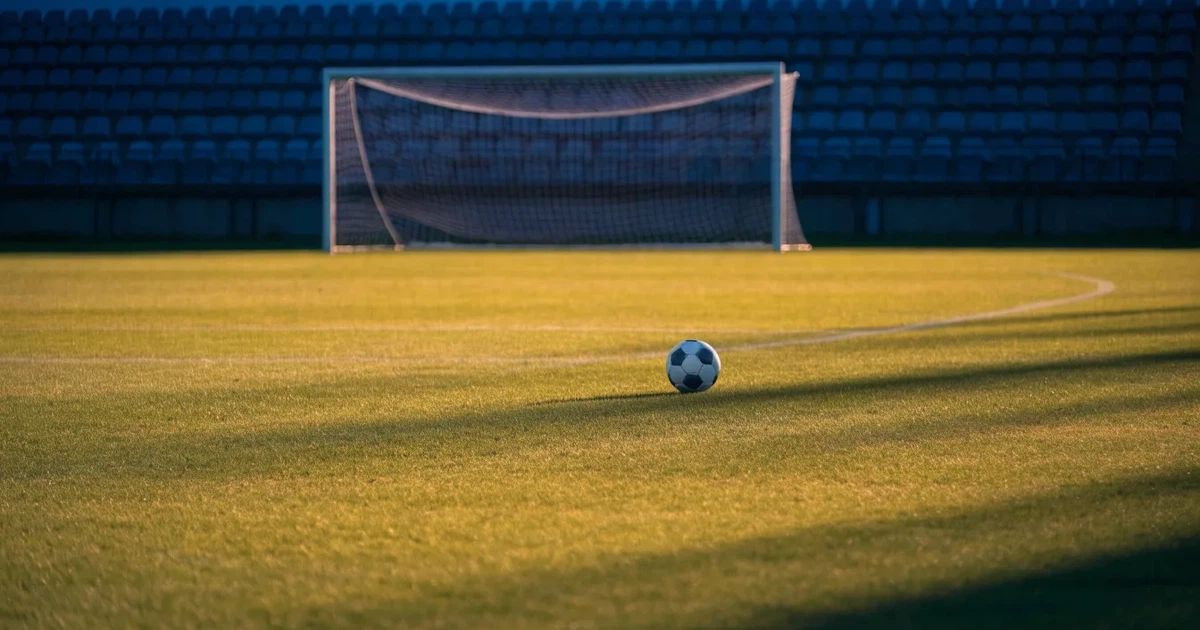 Ein Fußballplatz mit frisch gemähtem Rasen im Abendlicht, ein Ball liegt im Strafraum, im Hintergrund unscharf eine leere Tribüne.