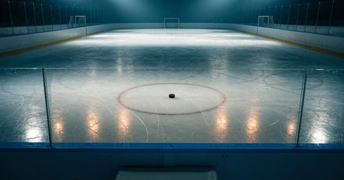 Eine leere Eisfläche in einer Halle mit Banden und Flutlicht, auf dem Eis liegt ein einzelner Puck.