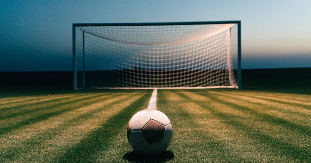 Ein Fußballtor mit Netz im Gegenlicht auf einem leeren Platz am Abend, davor ein Ball auf dem Rasen.